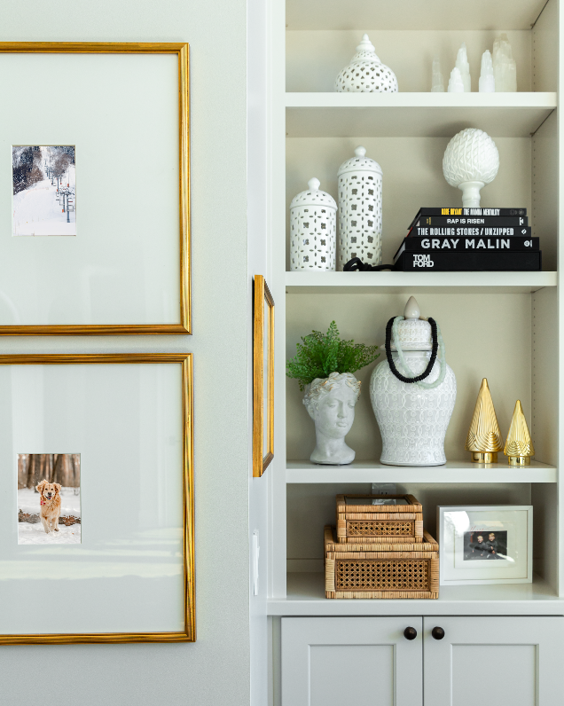 Stylish built-in shelving unit displaying a variety of decorative objects. The shelves house a collection of white and black patterned ceramic jars, assorted books on art and music, a bust planter filled with greenery, and sleek golden ornaments. Wicker boxes and framed photographs add a personal touch to the setup, all against a soft gray background that complements the home's refined style.